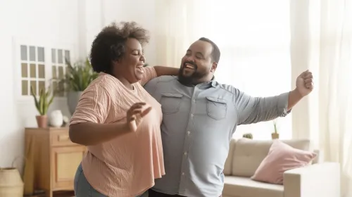 A joyful mature couple holding hands and dancing at home, representing consistency in physical activity and overall wellbeing during weight management.